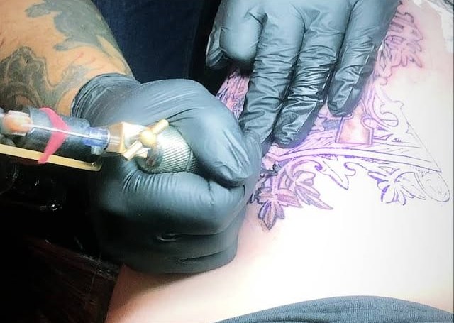 Tatuoija työssä: kädet mustissa käsineissä ja tatuointikone ihon päällä, luonnos näkyy violettina.